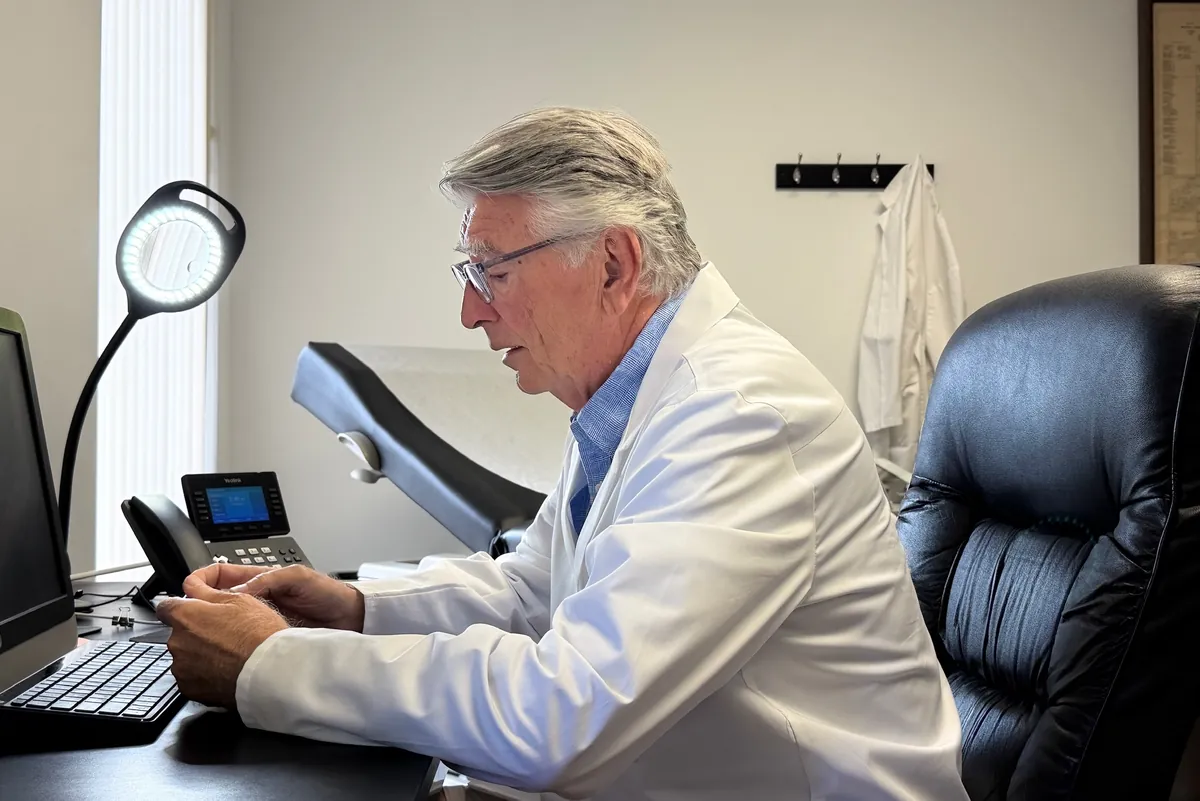Dr. Carroll at his desk computer, with a desk phone and an exam table beside him