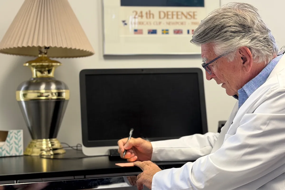 Dr. Carroll writing at his desk in the Maitland office, a lamp and a framed sailing poster behind him