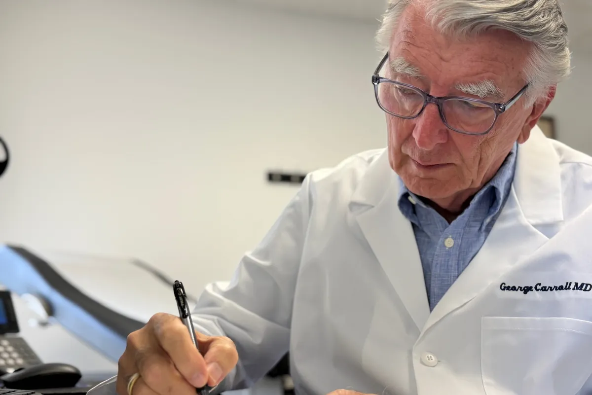 Dr. Carroll writing at his desk in a white coat, with an exam table behind him