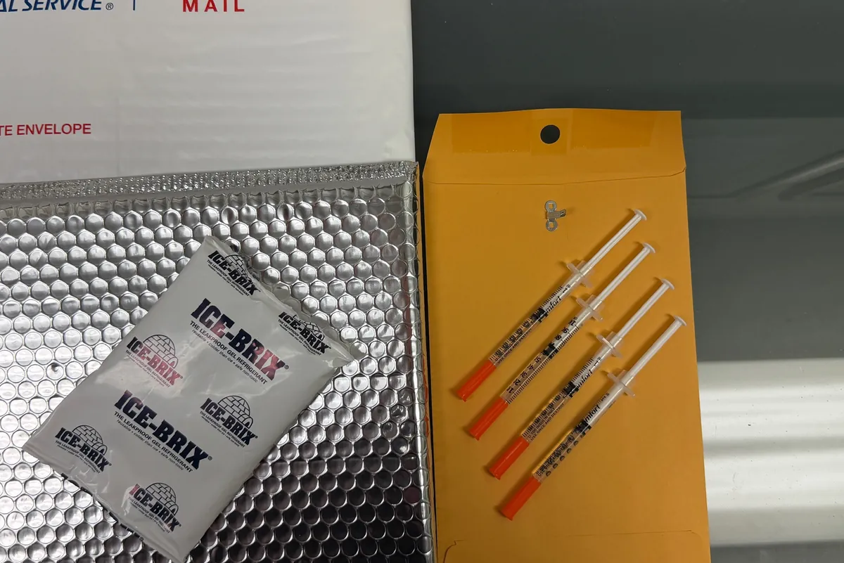 A shipping kit laid out on a table: a Priority Mail envelope, an insulated mailer, a cold pack, and four syringes on a paper envelope