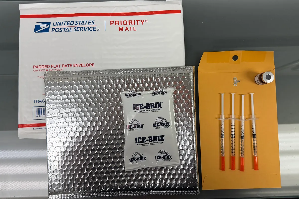 A shipping kit laid out on a table: a Priority Mail envelope, an insulated mailer with a cold pack, and four syringes and a vial on a paper envelope